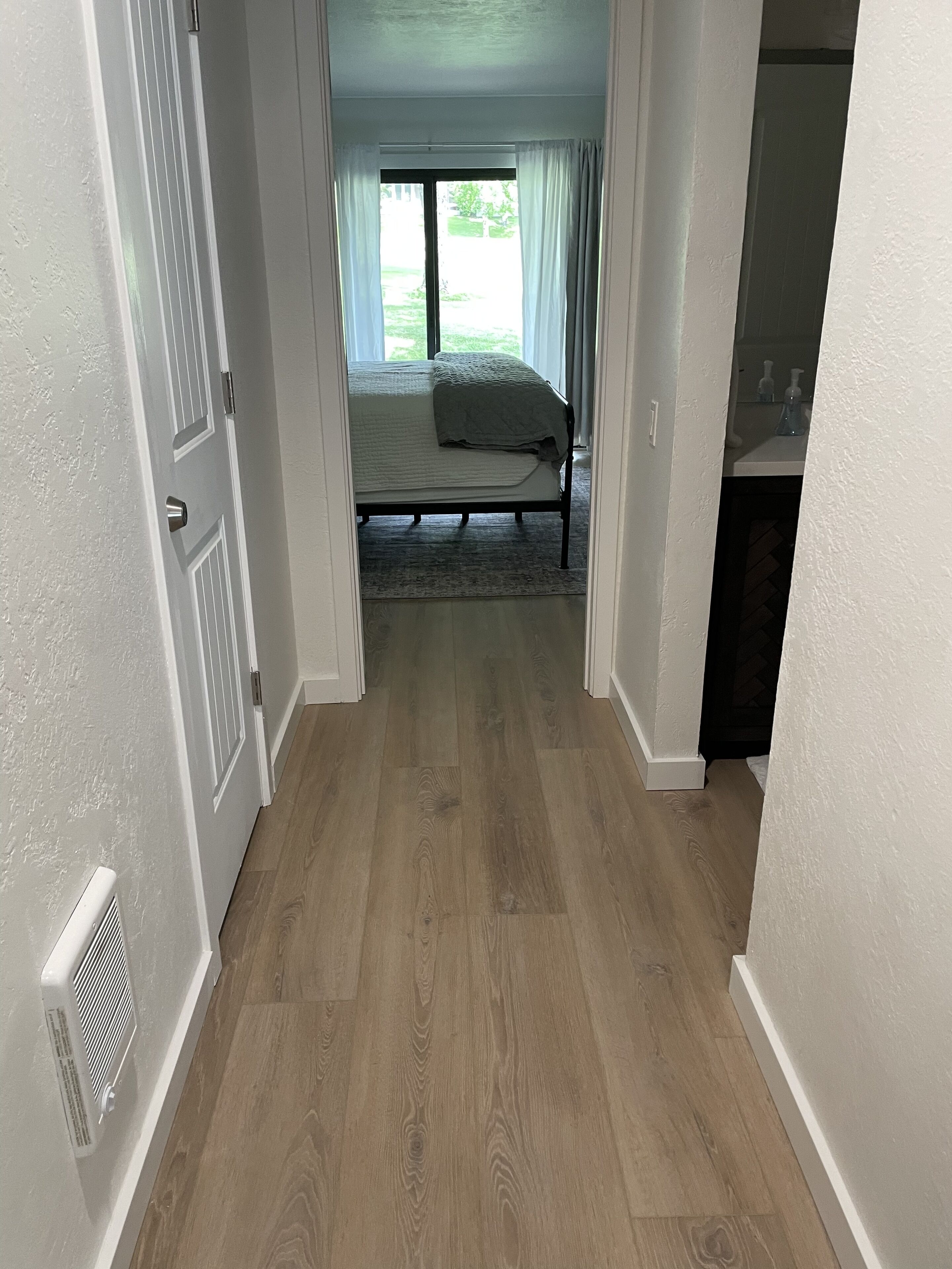 A hallway leading to a bedroom with a bed, a radiator, and a view of a garden through a window.