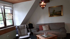 Un salon chaleureux avec un plafond haut, un plafond en bois, un canapé gris agrémenté de coussins rouges et beiges, une table basse en bois et un fauteuil avec un coussin.