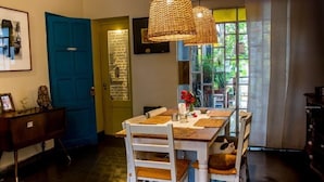 Un comedor con una mesa de madera y sillas blancas, una puerta azul y una lámpara colgante de mimbre.