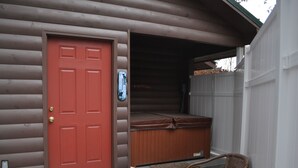 A log cabin with a red door, a hot tub, and a wicker chair outside.
