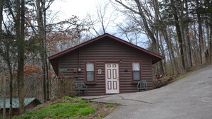 A small log cabin with a red roof, a white door, and two windows with shutters. There are two green chairs outside.