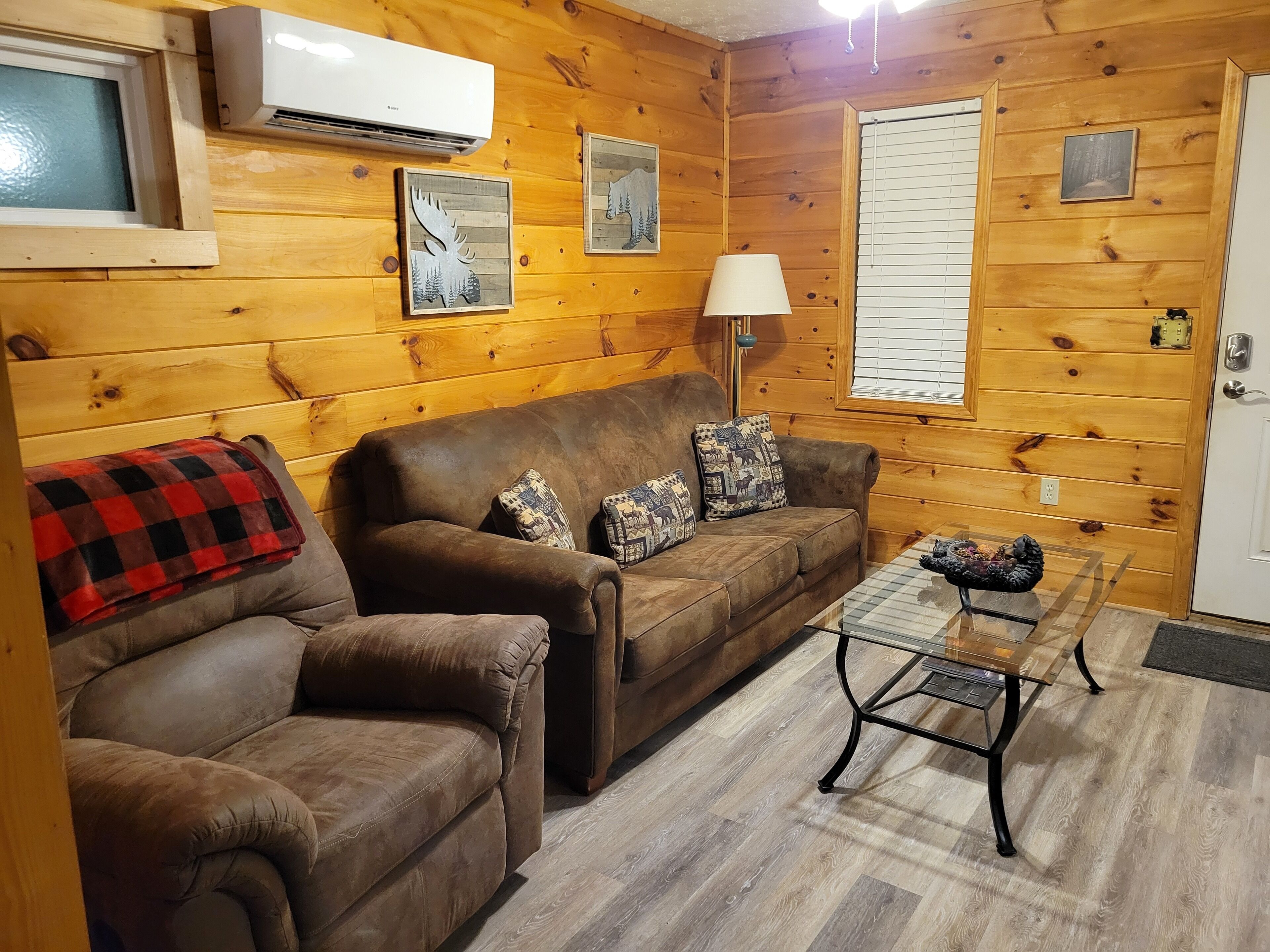 A cozy living room with a brown leather sofa, a matching armchair, and a glass coffee table. There are two wall-mounted air conditioners and framed pictures on the walls.