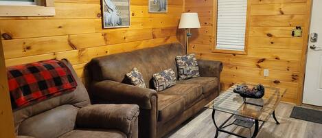 A cozy living room with a brown leather sofa, a matching armchair, and a glass coffee table. There are two wall-mounted air conditioners and framed pictures on the walls.
