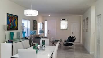 A modern dining area with a white table, chairs, and a chandelier. There is a sofa and a coffee table in the background.