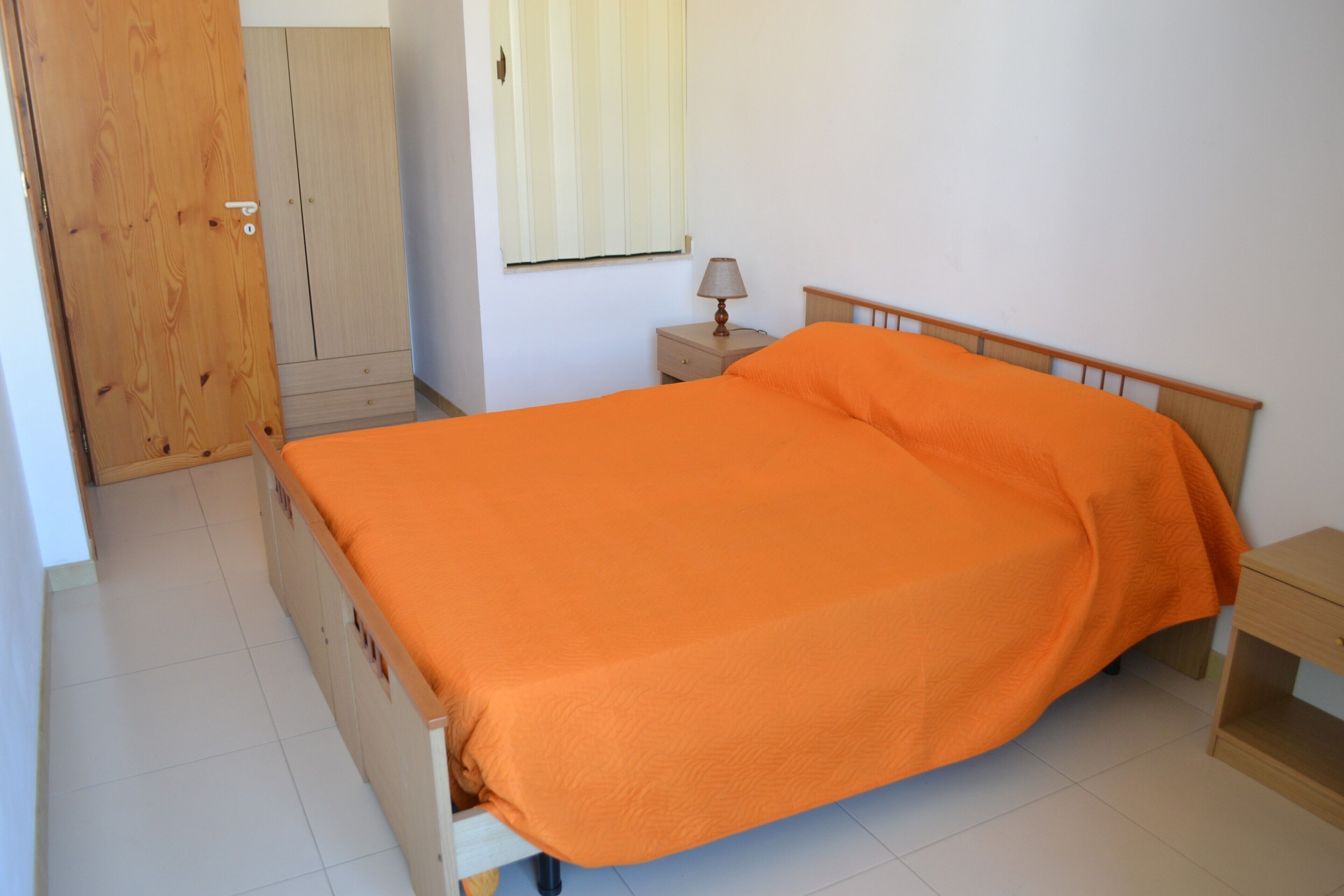 Quarto com cama, mesa de cabeceira, guarda-roupa de madeira e porta.