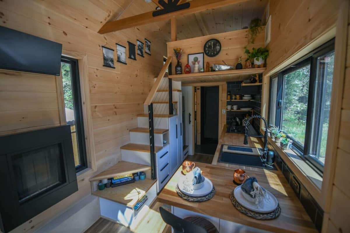 A compact wooden cabin interior with a staircase, a dining area with a table set for two, and a kitchen area with a sink and cabinets.