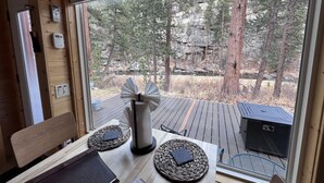 A wooden interior with a table set for two, a window with a view of a forest, and a grill outside. - The Woolly Bugger Inn (Drake)