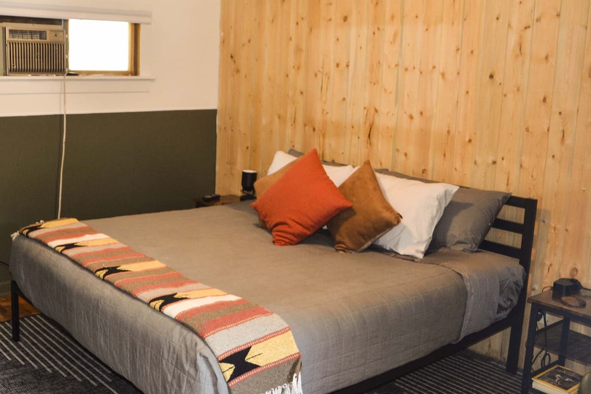 A bed with a grey bedspread, a colorful blanket, and two pillows.