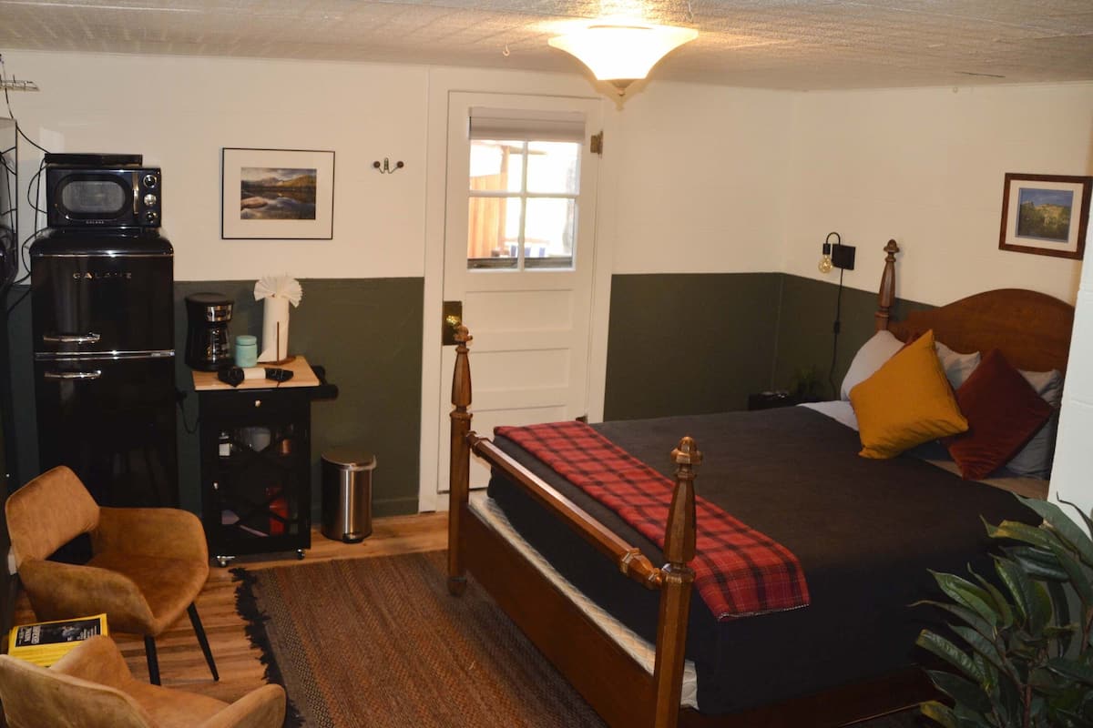 A bedroom with a bed, a wooden chair, a desk with a microwave, a coffee maker, and a small trash bin.