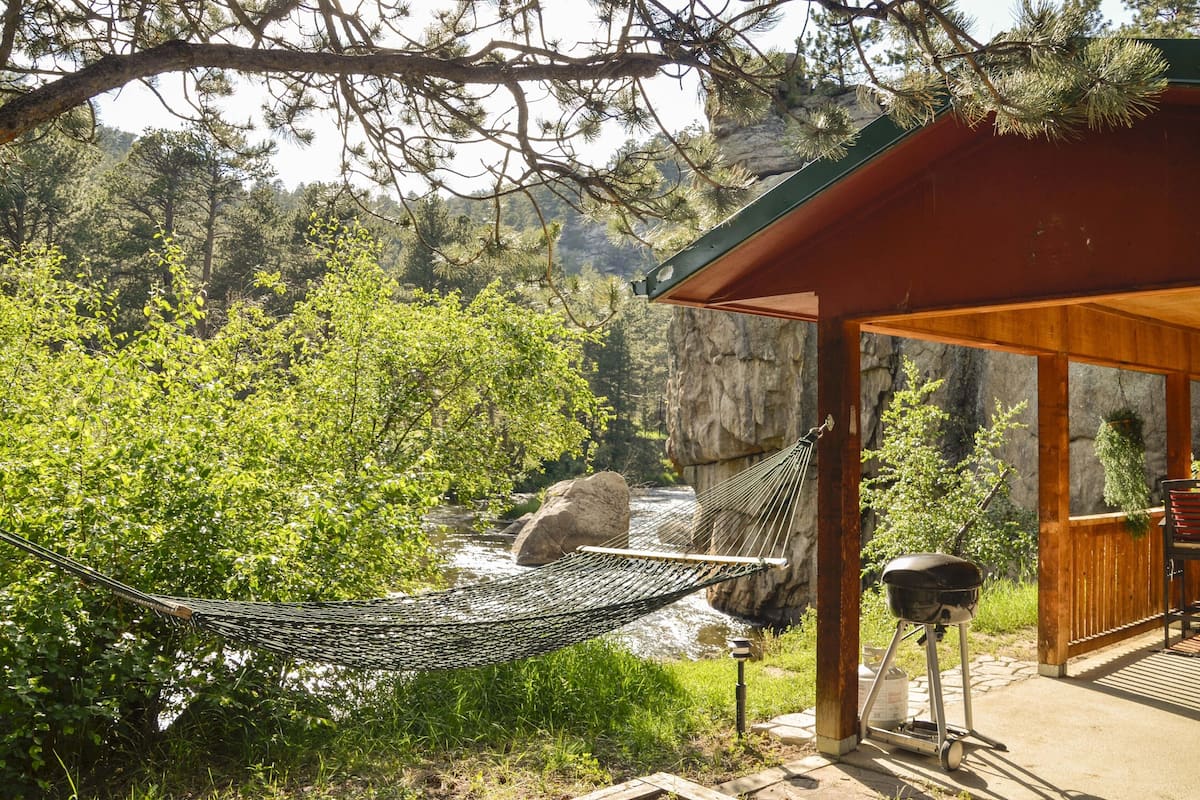 A hammock tied to a wooden structure overlooking a river, with a grill and seating area nearby.