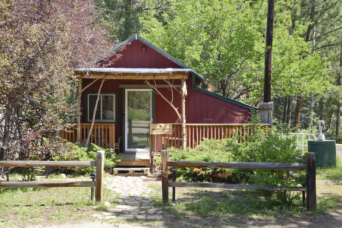 A small cabin with a wooden deck and a porch, surrounded by greenery.