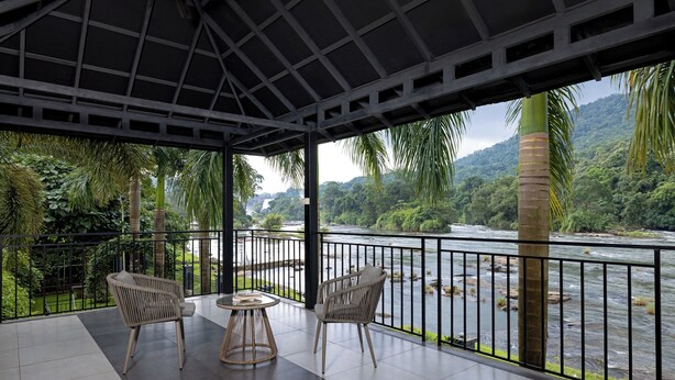A covered outdoor seating area with two wicker chairs and a small round table, overlooking a river and lush greenery. - Casa Rio Resort Athirapilly (Chalakudy)