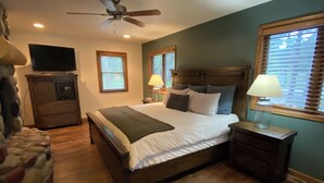 Un dormitorio con cama, cabecera de madera, mesitas de noche con lámparas, ventilador de techo y televisor sobre un aparador.
