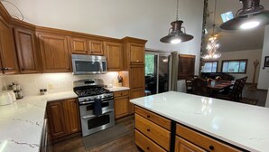 Una cocina con armarios de madera, una estufa de acero inoxidable, un microondas y un refrigerador. Se ve un área de comedor con una mesa y sillas a través de una puerta abierta.