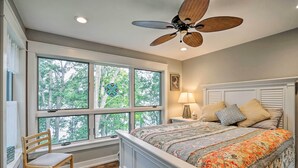 Una camera da letto con una grande finestra, un letto con copriletto floreale, una sedia in legno, un comodino con una lampada e una ventola a soffitto.