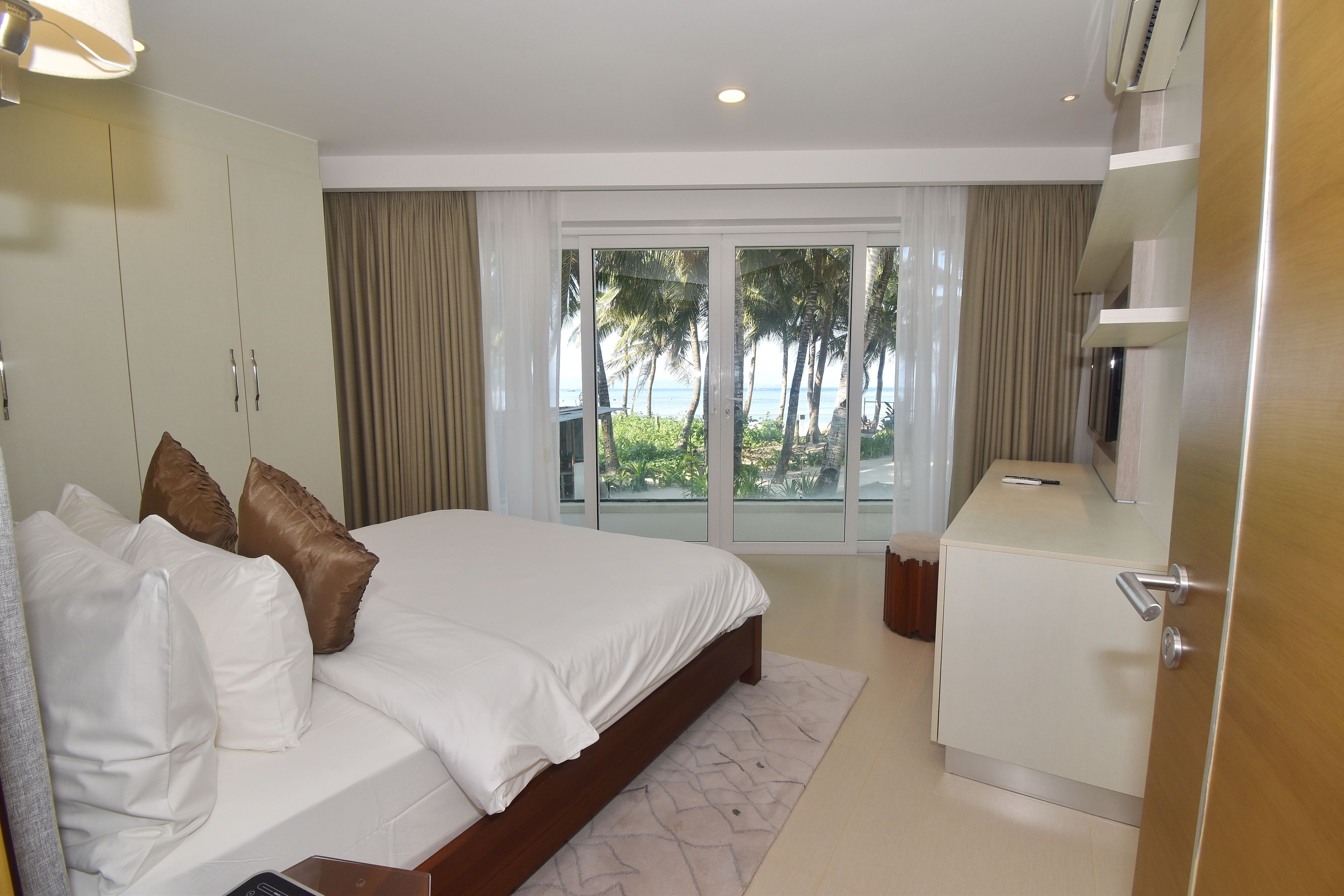 A hotel room with a large bed, a wardrobe, a desk, and a view of palm trees and the ocean through the sliding glass doors.