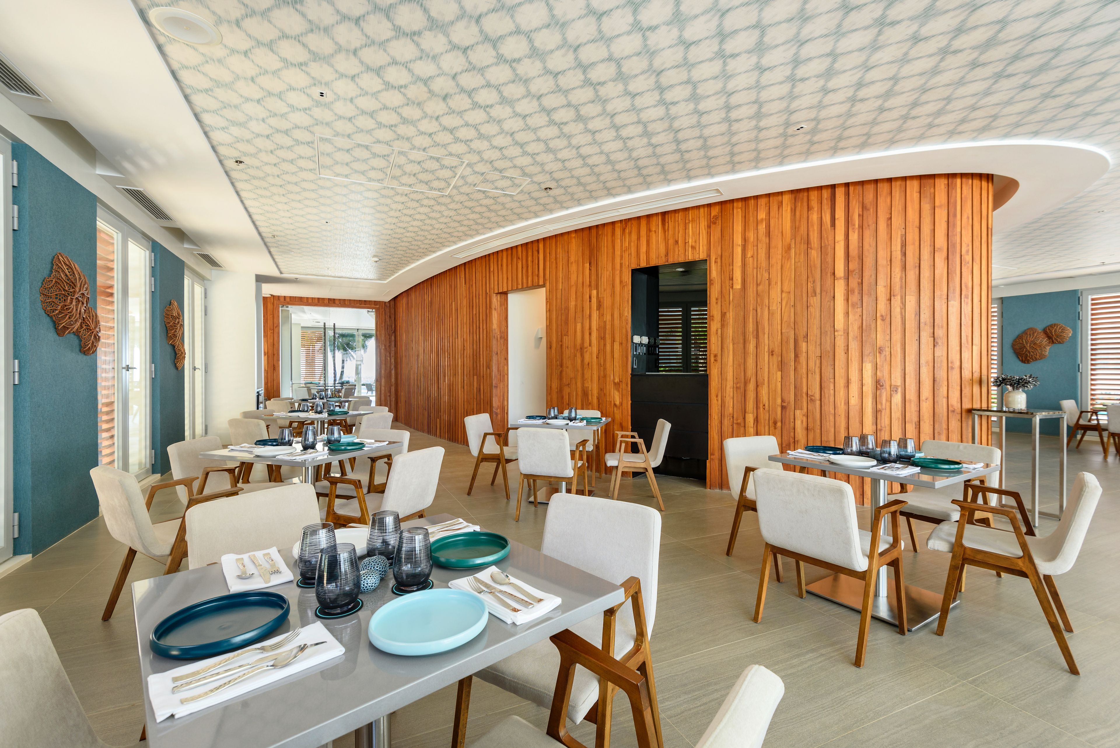 A modern dining area with wooden paneling and a patterned ceiling. Tables are set with plates, glasses, and cutlery.