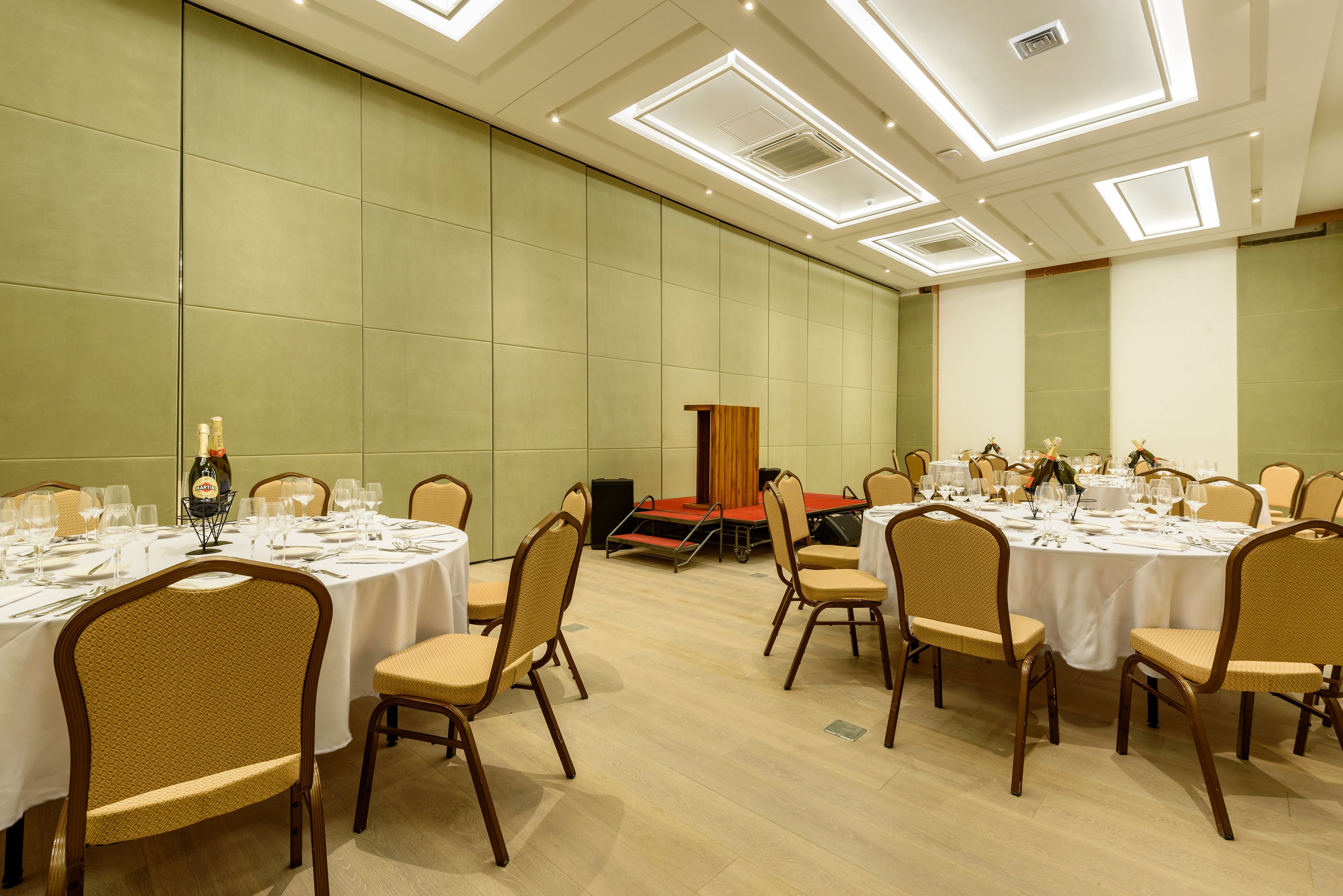 Un salón de conferencias con varias mesas dispuestas para una reunión, equipado con sillas y mesas, y un área de escenario al fondo.