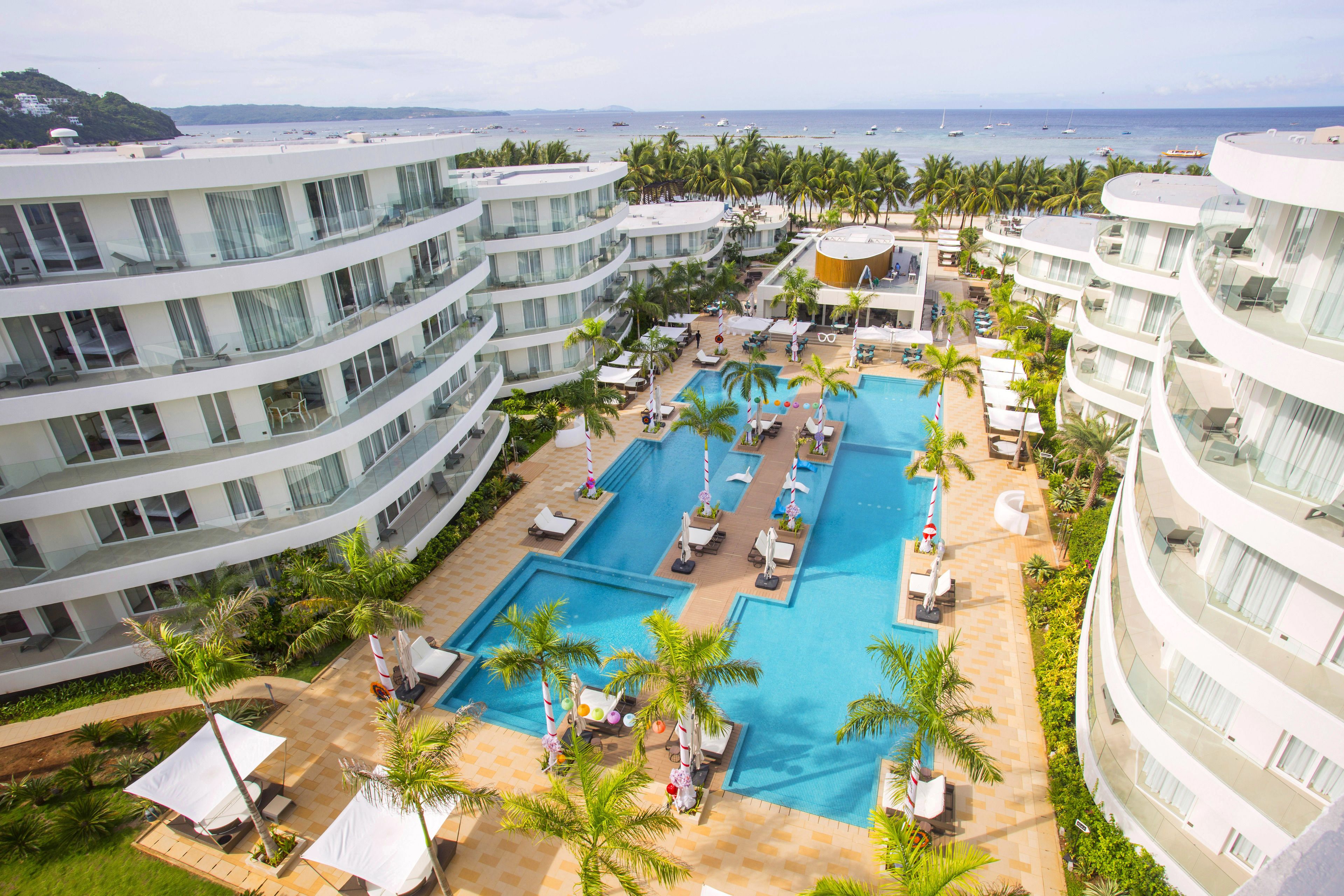 A hotel with multiple floors, a central swimming pool surrounded by sun loungers and palm trees, and a view of the sea.
