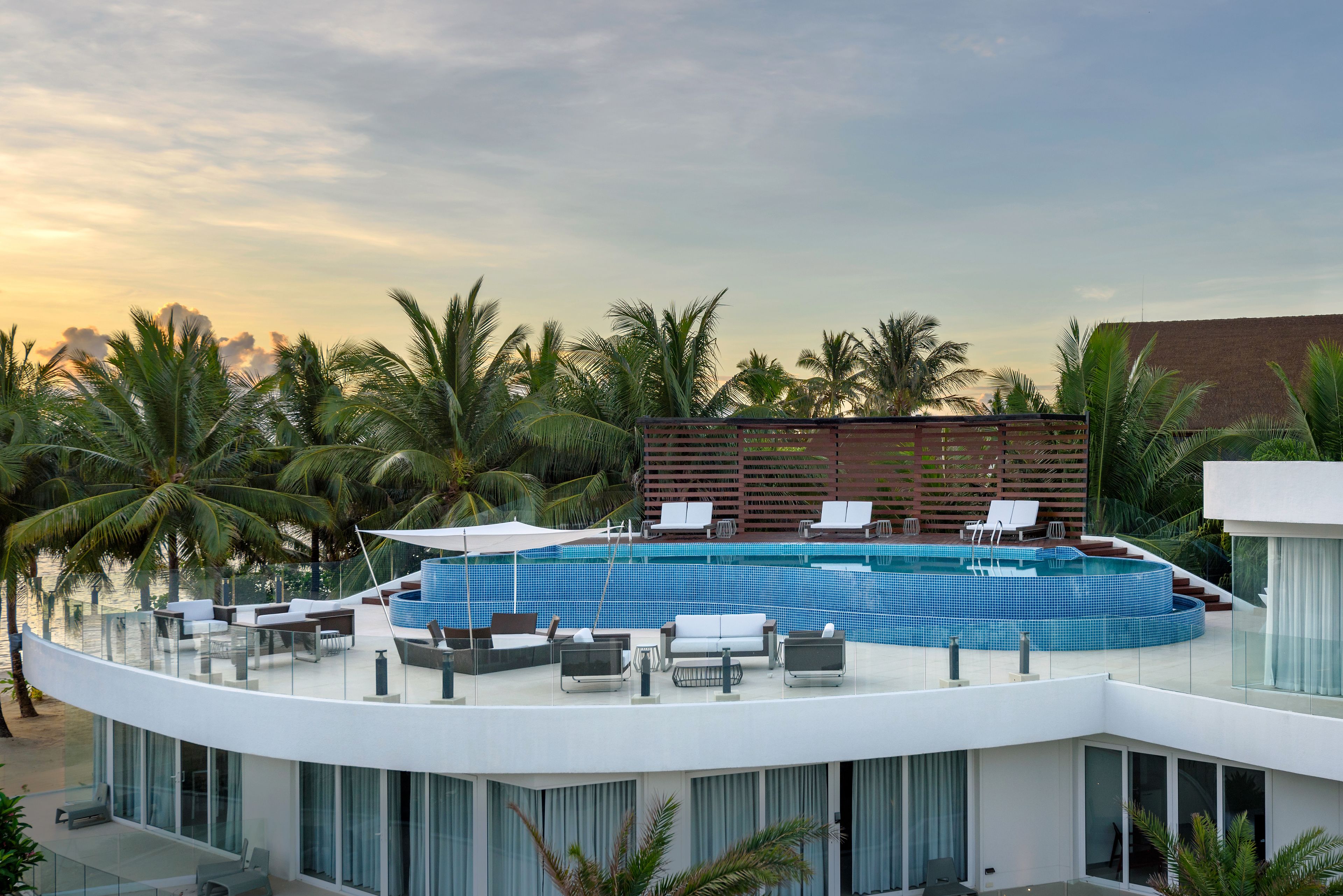 A modern hotel with a rooftop infinity pool, lounge chairs, and palm trees.