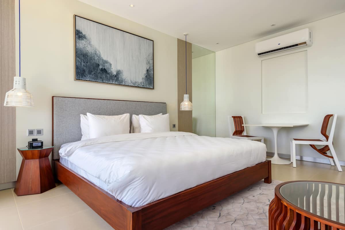 Una habitación de hotel moderna con una cama grande, una mesita de noche, una silla, una pequeña mesa y un aire acondicionado de pared.