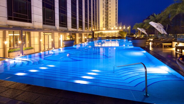 A swimming pool with a blue-lit design, surrounded by lounge chairs and umbrellas, with a high-rise building in the background.