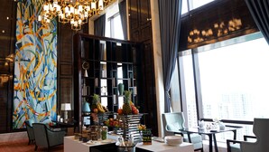 A modern hotel lobby with a large abstract painting, a chandelier, a fruit display, and a view of the city through large windows.