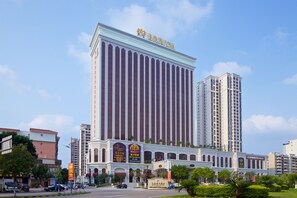 A large hotel with a prominent vertical facade and arched windows, surrounded by other buildings and greenery. - Mingzuo Hotel Zhongshan (Zhongshan)