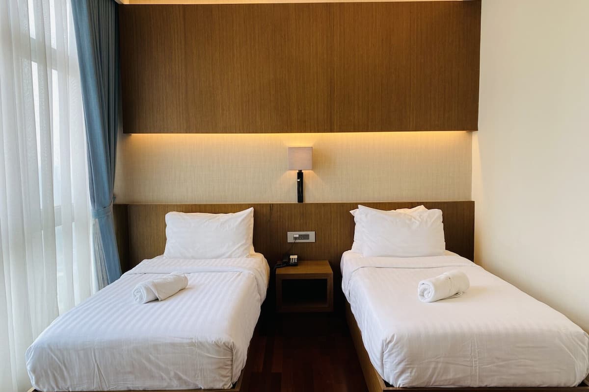 Une chambre d’hôtel avec deux lits, des têtes de lit en bois, une table de chevet, une lampe et une fenêtre avec des rideaux.