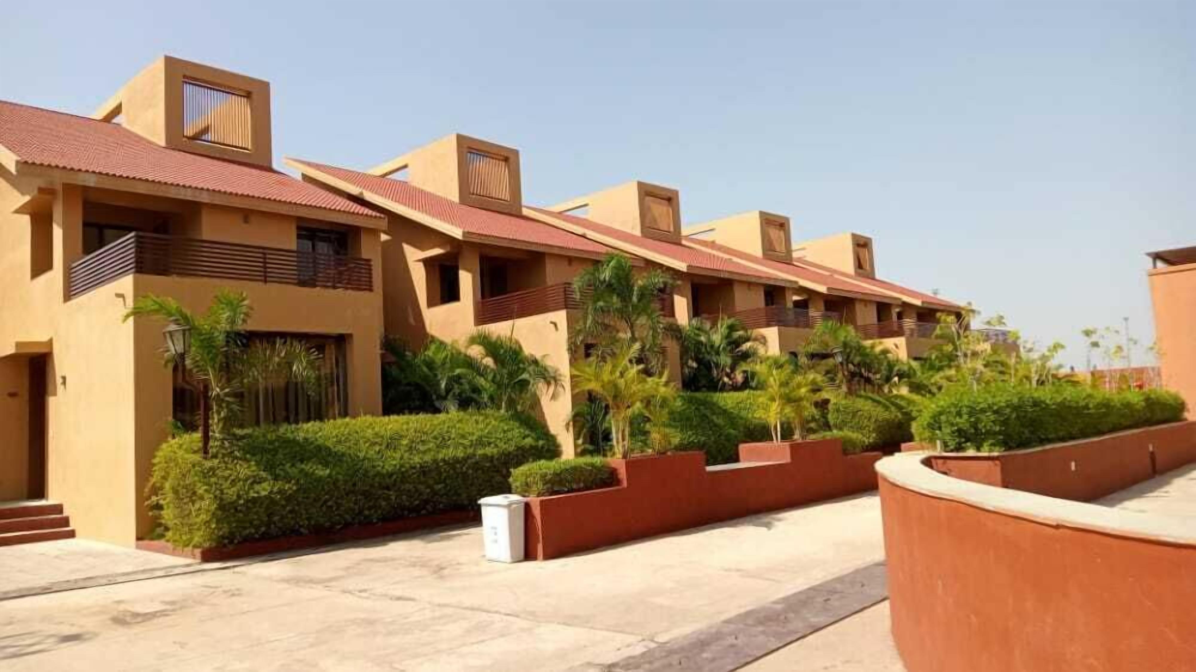 A row of modern, beige-colored buildings with flat roofs and balconies, surrounded by well-maintained greenery and a clear sky.