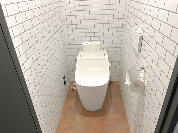 A small, white-tiled bathroom with a toilet, two rolls of toilet paper, and a towel dispenser.