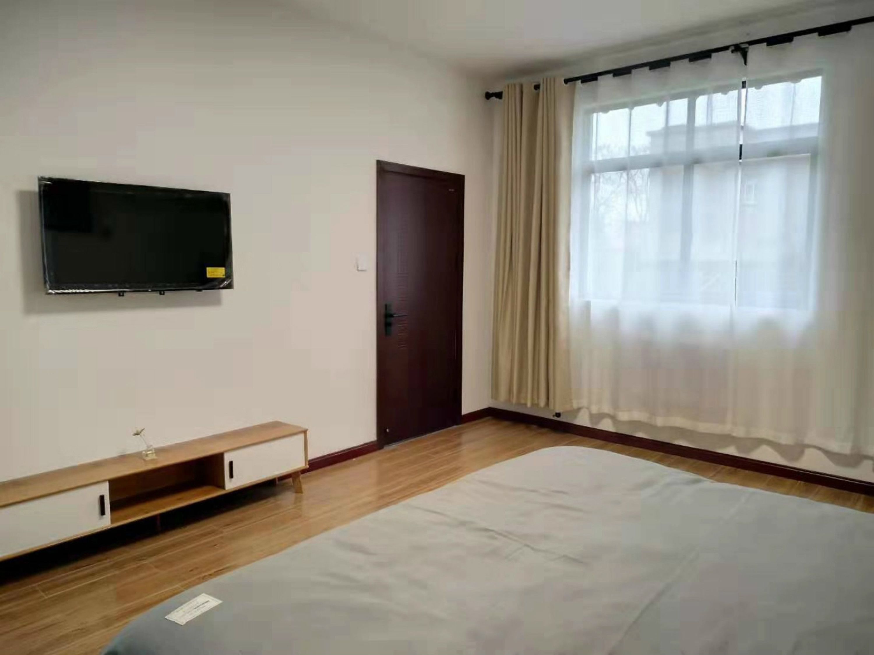 Quarto com cama, televisão na parede, porta e janela com cortinas.
