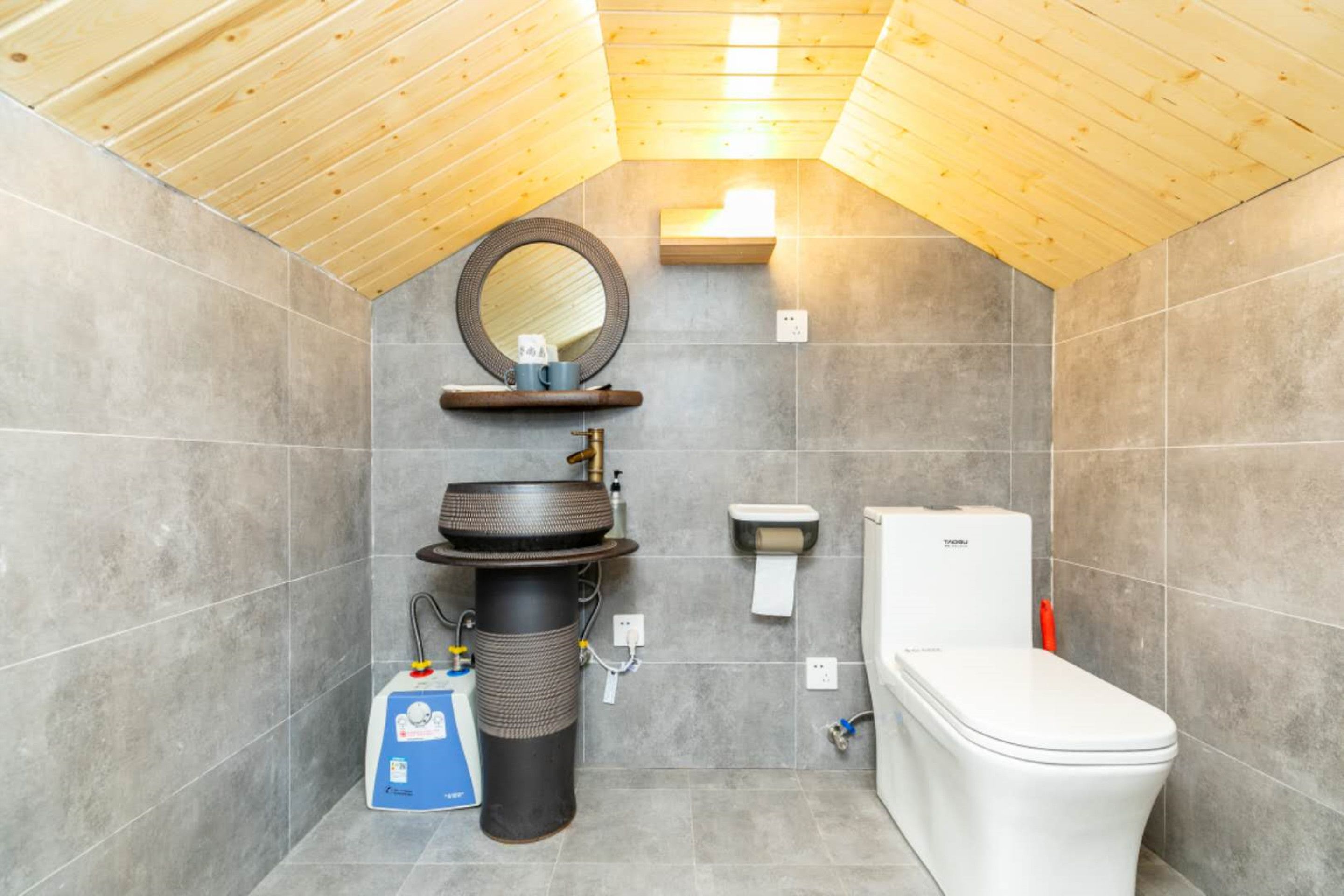Un baño pequeño con techo de madera, un espejo redondo, un lavabo, un inodoro y una pequeña repisa con artículos de aseo.