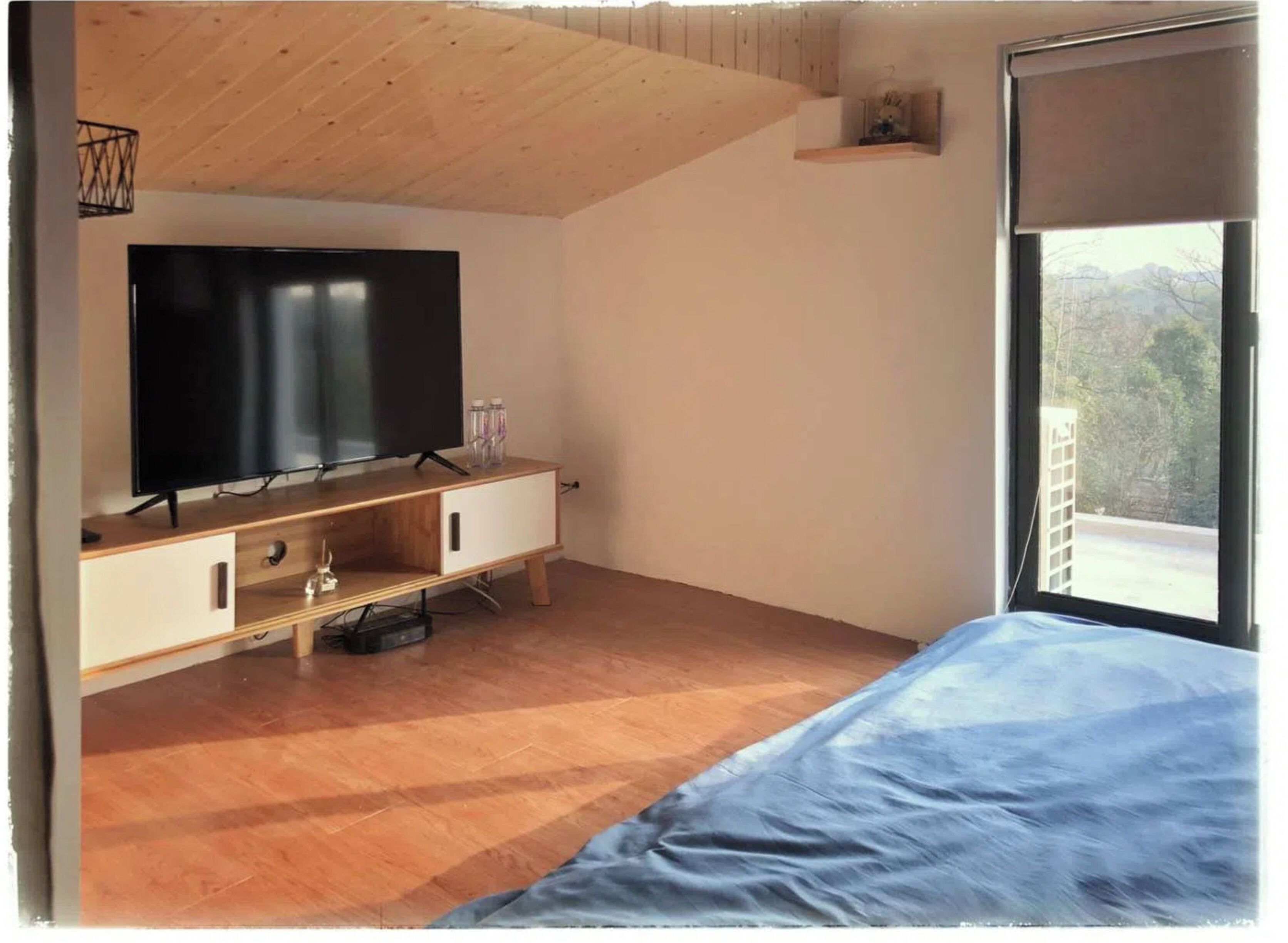 Un dormitorio con techo de madera, un televisor sobre un soporte de madera, una cama con cubrecama azul y una ventana con vista a árboles.
