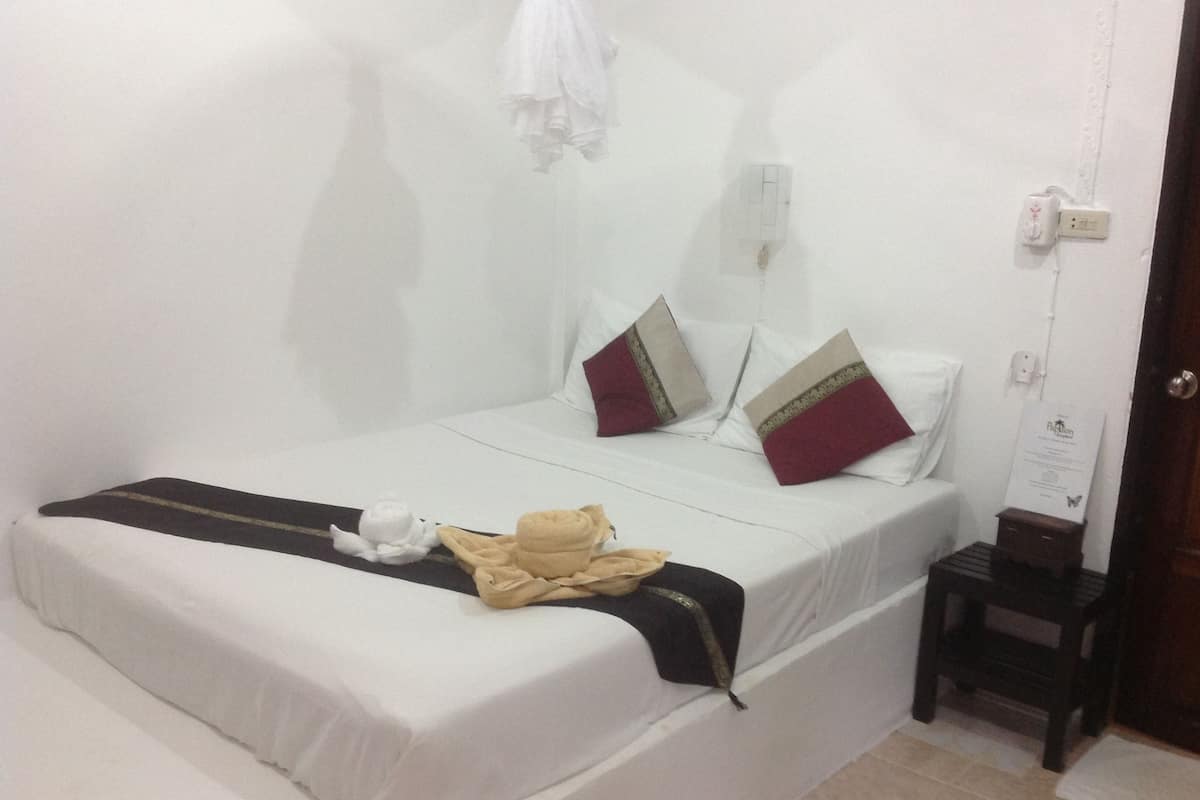 A neatly made bed with white bedding and two decorative pillows, a small bedside table with a lamp, and a wall-mounted power outlet.