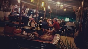 A dimly lit indoor space with tables and chairs, hanging lanterns, and a bar area in the background. - Papillon Bungalows (Ko Lanta)