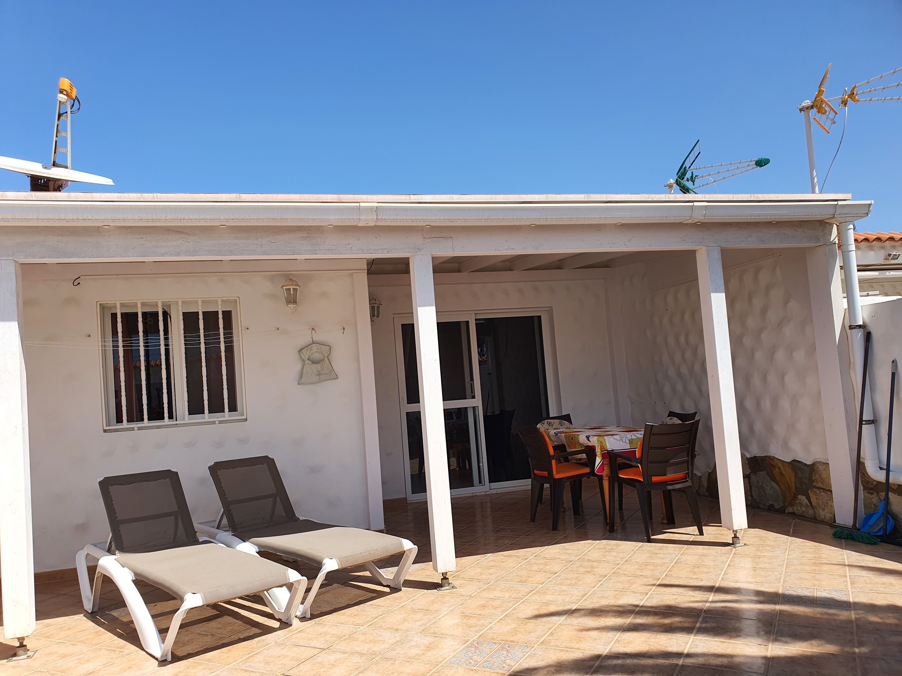 Eine überdachte Terrasse mit zwei Liegestühlen, einem Tisch mit Stühlen und einem Fenster mit Gitterstäben.