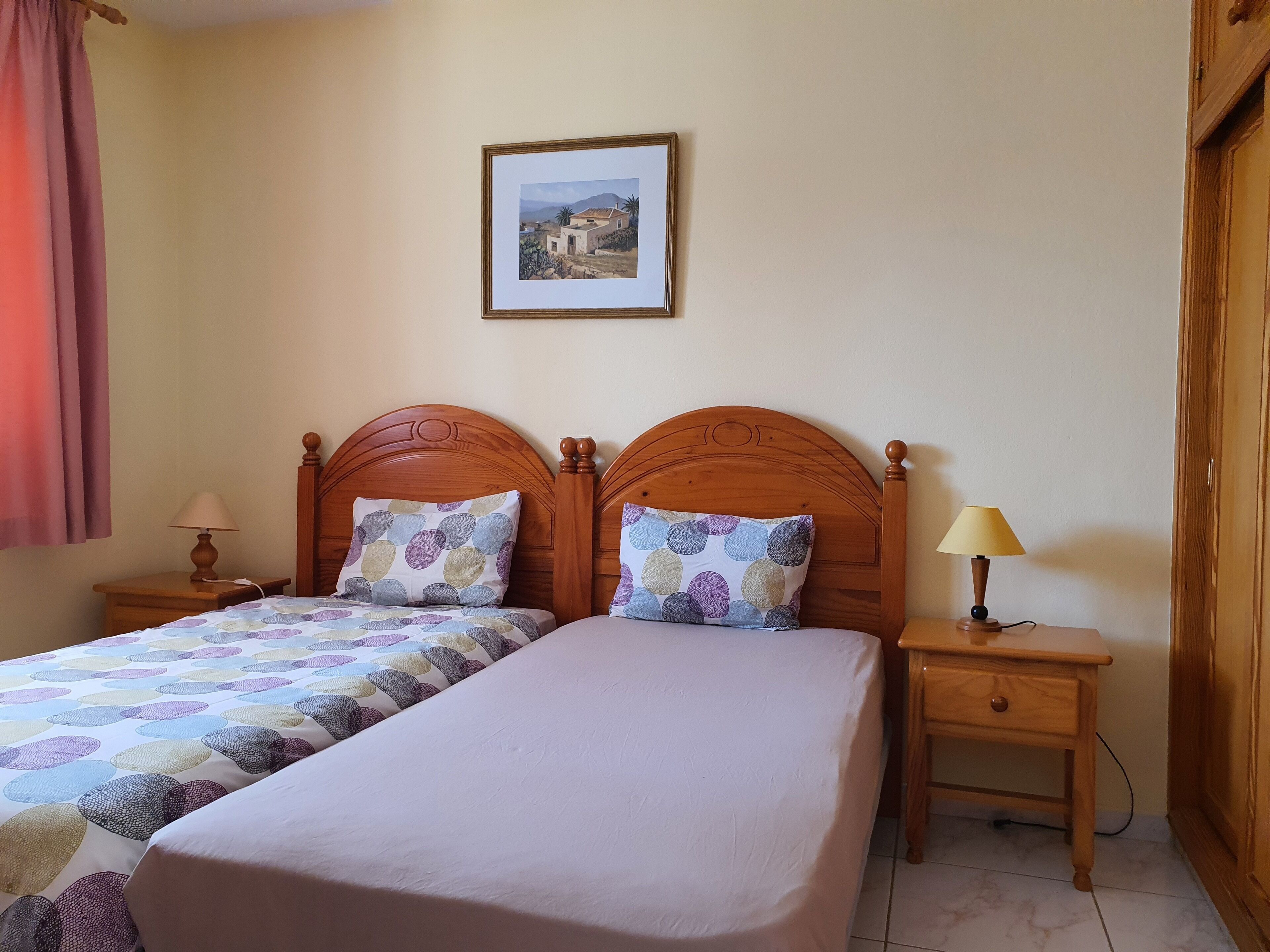 A double bed with a patterned bedspread, two pillows, a nightstand with a lamp, and a framed picture on the wall.