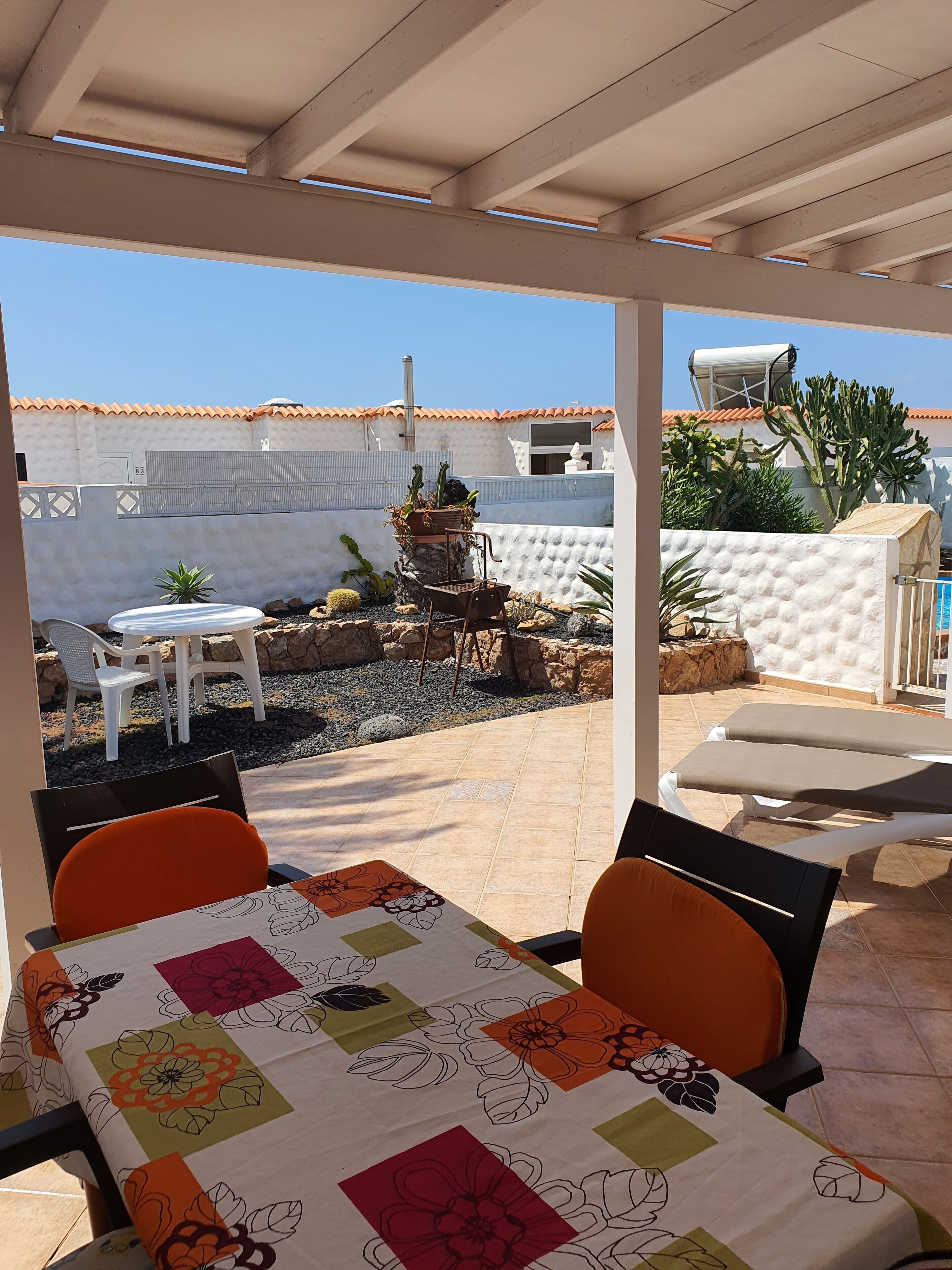 An outdoor patio area with a table set for dining, surrounded by chairs. There are potted plants and a small table with a chair nearby. The patio is enclosed by a white wall with a tiled roof.