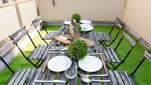Un espace extérieur rectangulaire aménagé pour les repas, avec des tables et des bancs en bois, entouré d’un petit muret et d’une pelouse.
