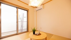 Une pièce dotée d’un sol recouvert de tatamis, d’une porte-fenêtre coulissante donnant sur un balcon, d’une petite table en bois ornée d’une plante et d’un vase, et d’un luminaire fixé au plafond.