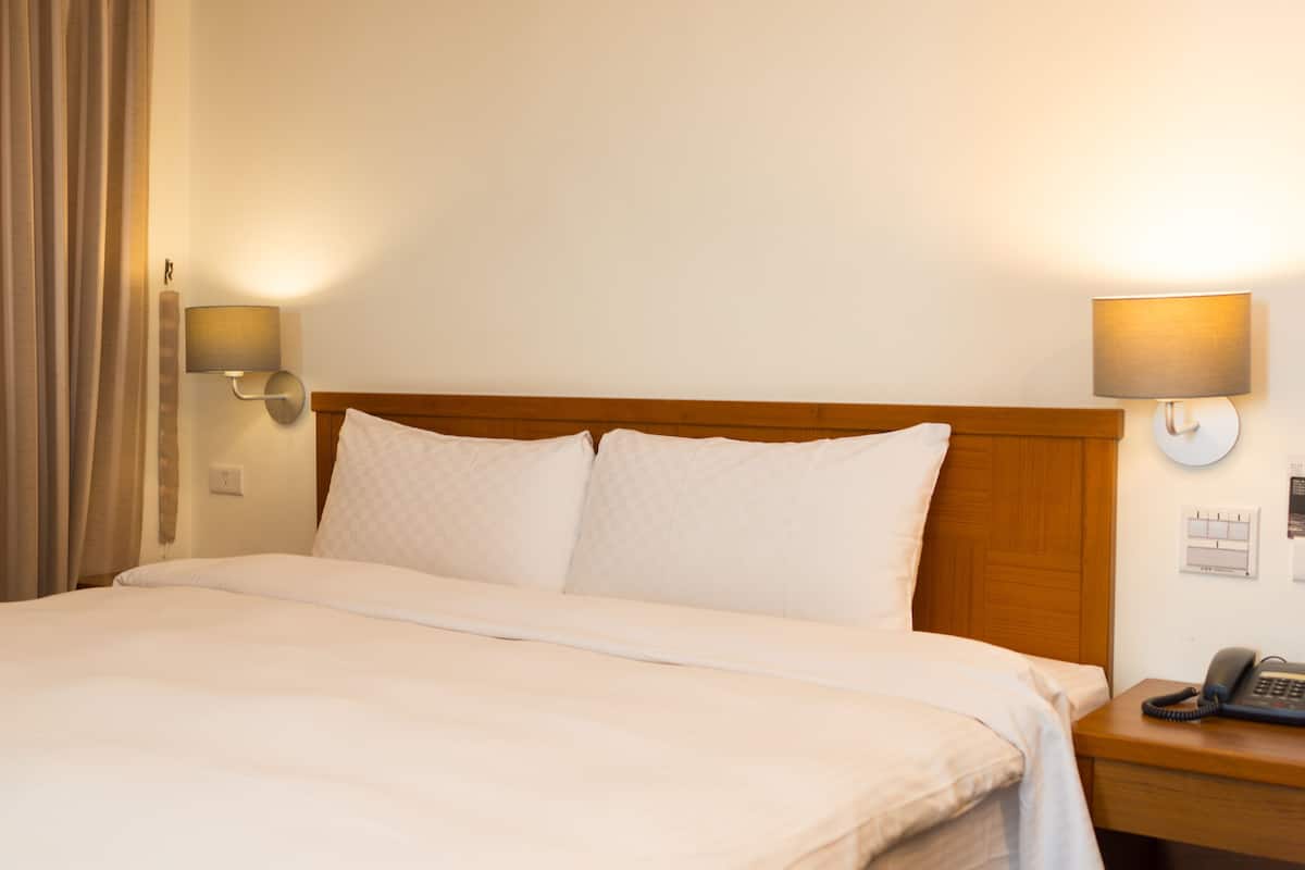 A hotel room with a wooden headboard, a bed with white linens, two wall-mounted lamps, a bedside telephone, and a wall calendar.