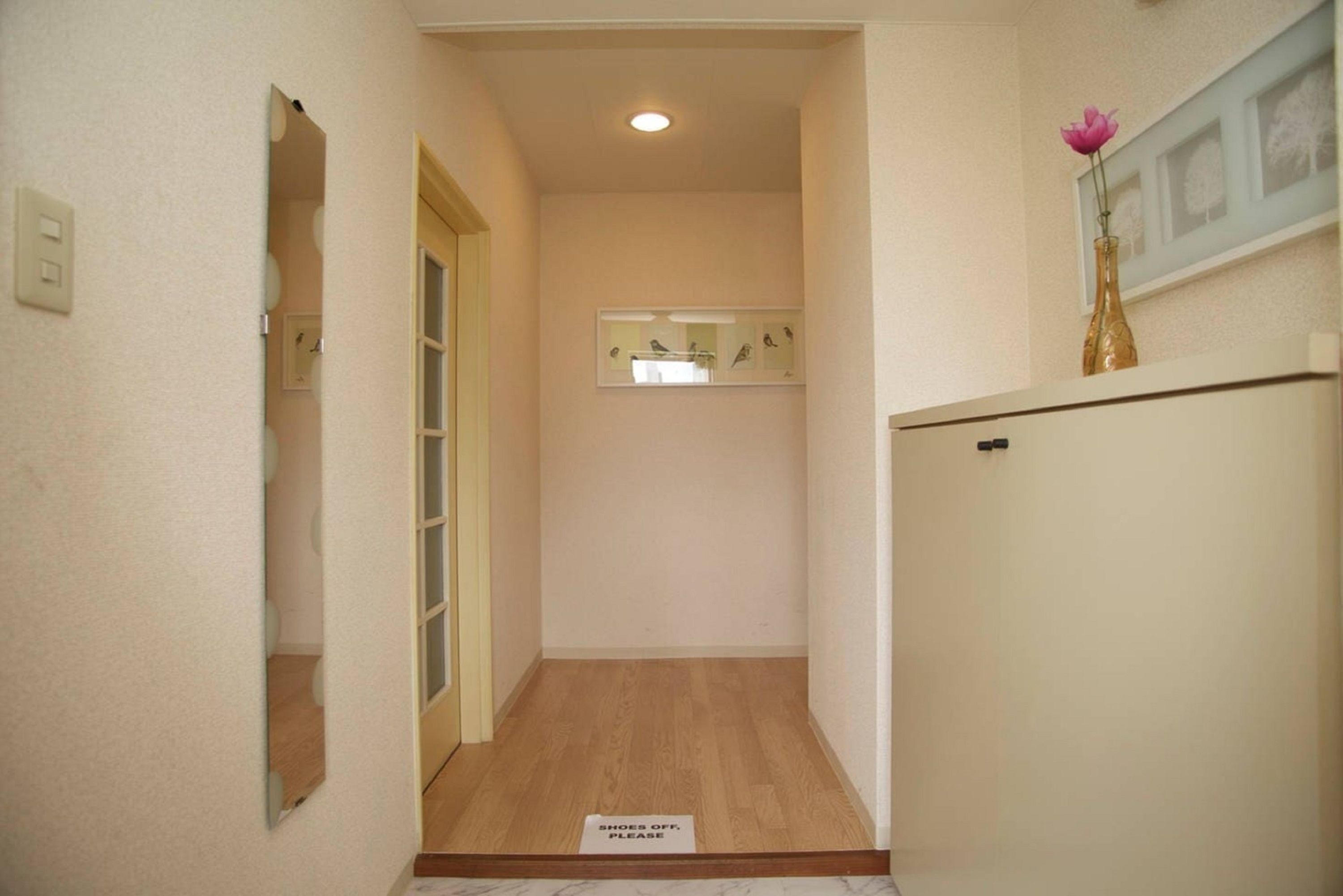 Un pasillo estrecho con suelo de madera, un cartel que dice 'Shoes Off, Please' en la entrada, un espejo a la izquierda y un jarrón con una flor rosa a la derecha.