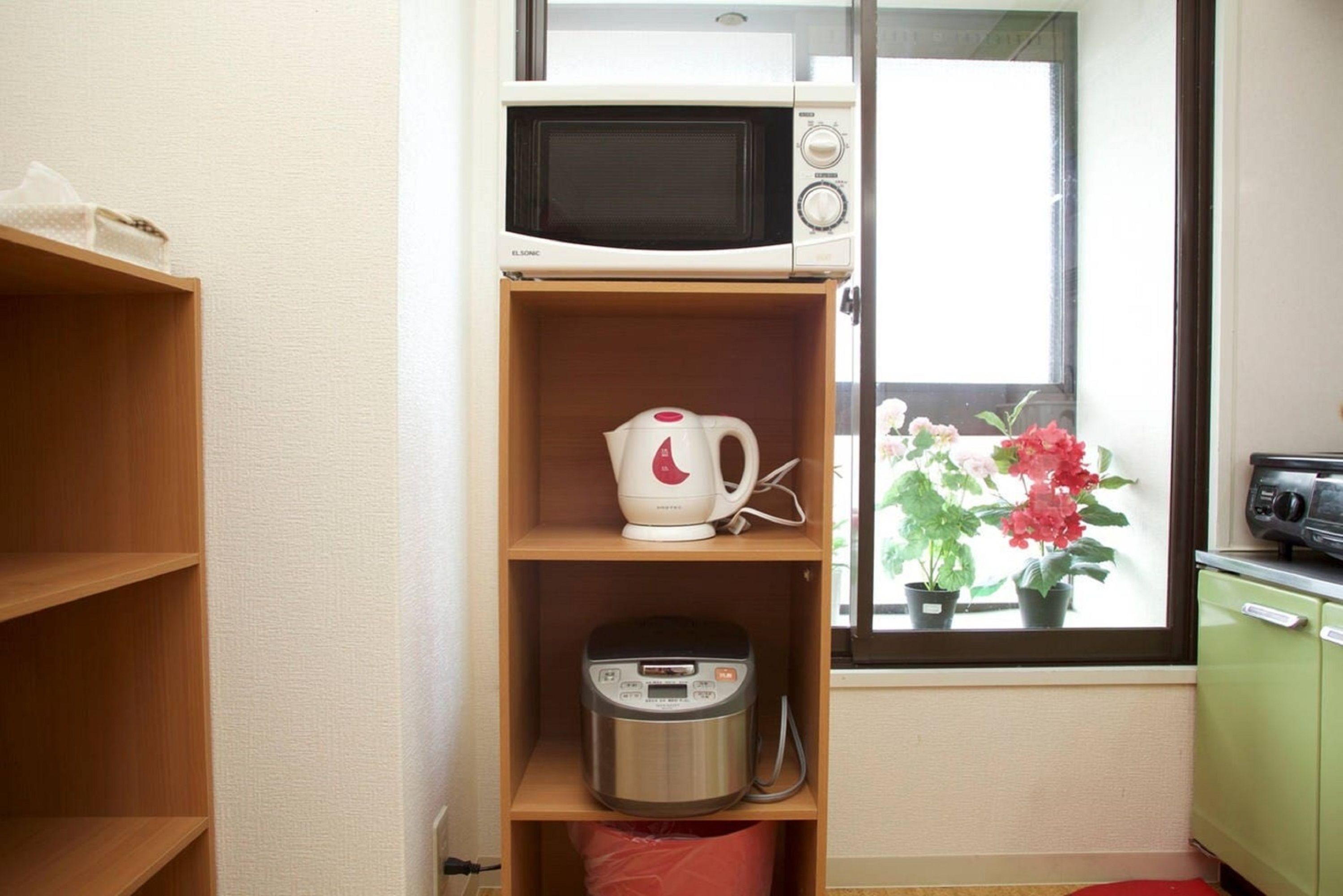 A compact kitchen with a microwave, a rice cooker, and a kettle on shelves, a window with potted plants, and a green cabinet.