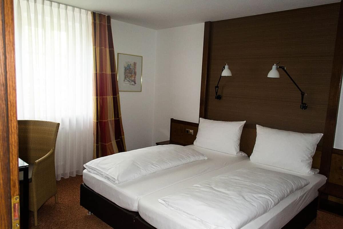 Un lit double avec du linge de lit blanc, une tête de lit en bois, deux lampes de chevet, une chaise en osier et une fenêtre avec des rideaux légers.