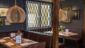 Un restaurant avec des tables et des banquettes en bois, des tableaux encadrés aux murs et une fenêtre ornée d’une grille métallique.