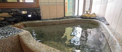Una bañera de piedra con superficie de agua cristalina, rodeada de paredes de piedra y suelo de baldosas.