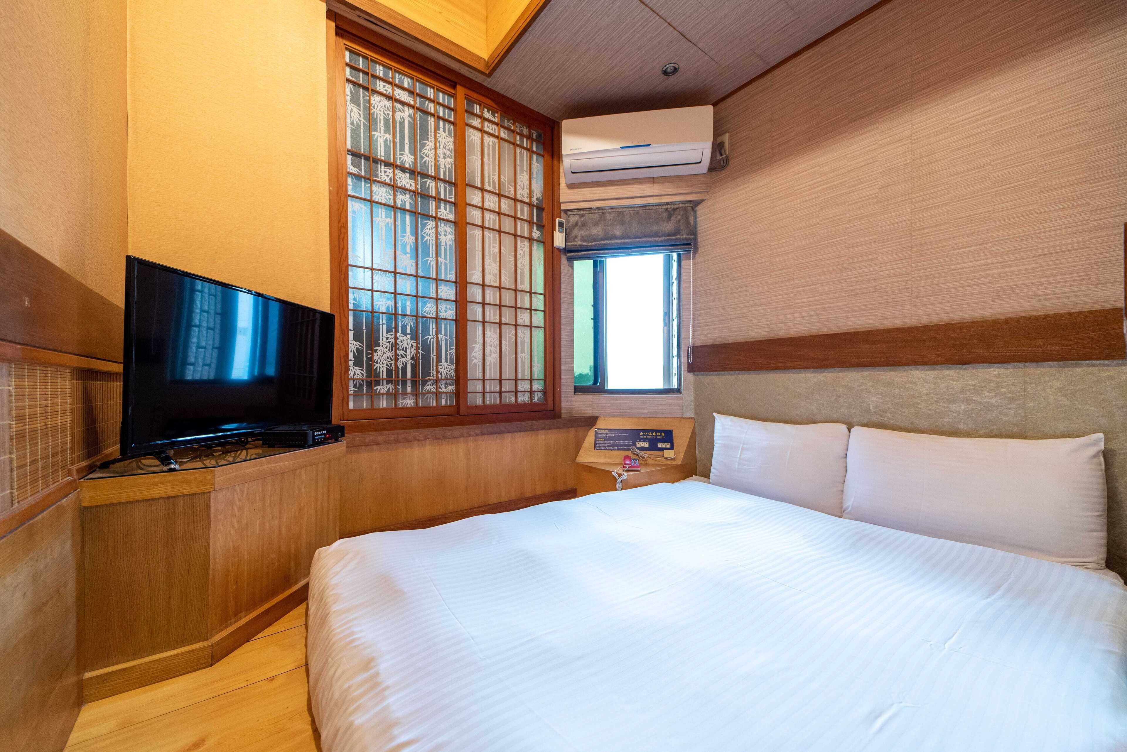 Ein Hotelzimmer mit einem großen Bett, einem Fernseher, einem Fenster mit traditioneller Gitterstruktur und einer Klimaanlage.
