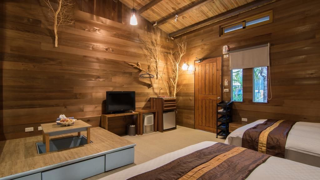 A wooden cabin-style room with a bed, a small table with a bowl of fruit, a television, and a window with a view of palm trees.