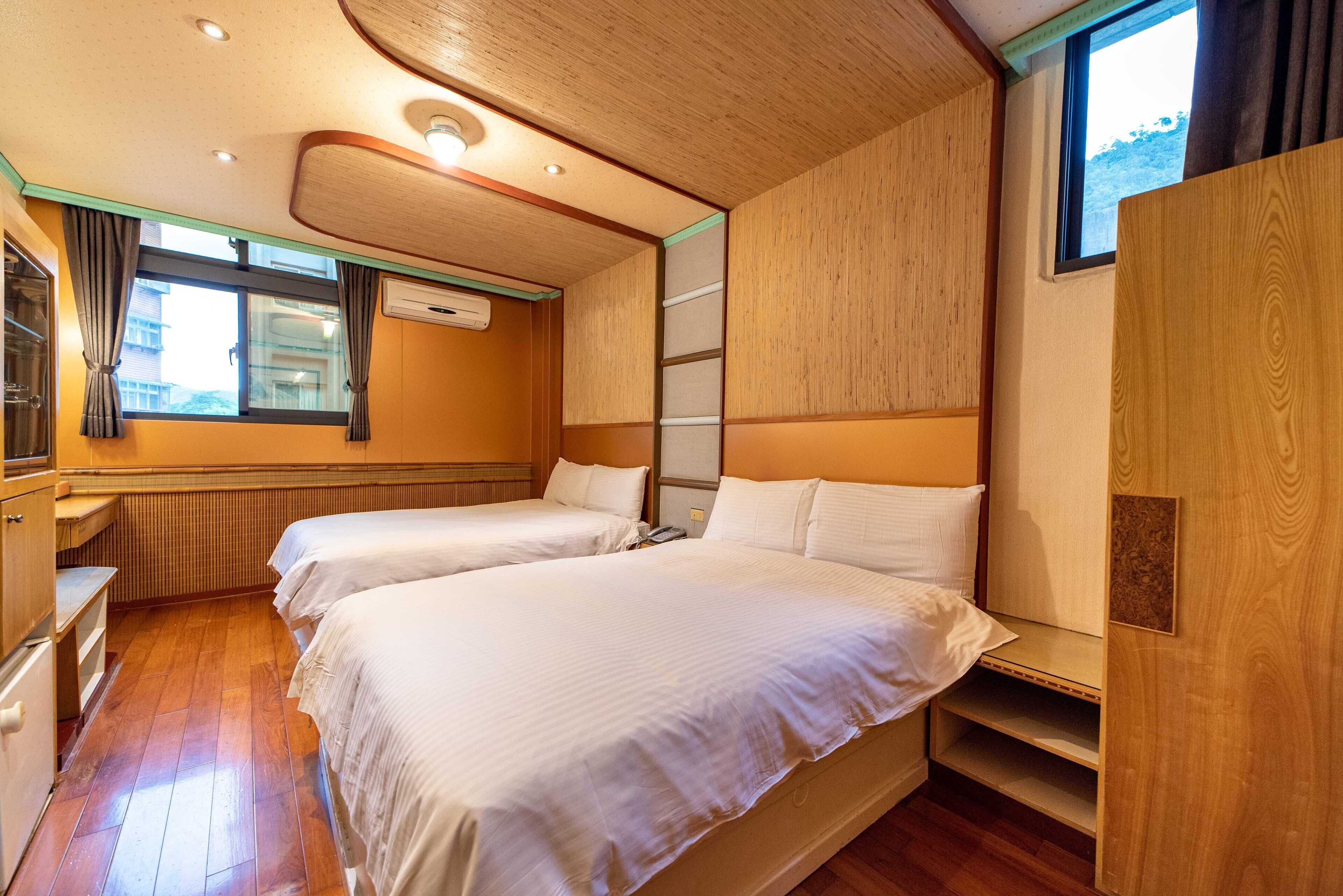 A room with two beds, wooden flooring, a window with curtains, and built-in shelves.
